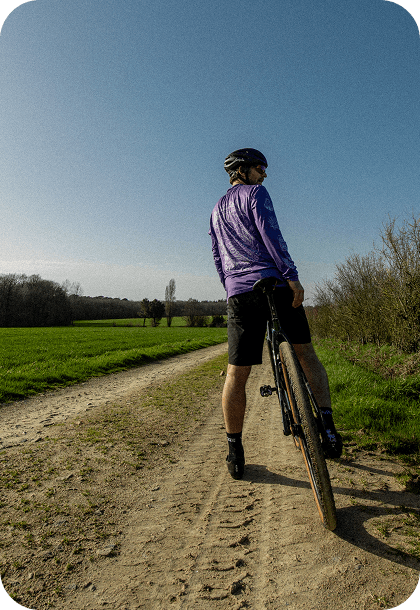 Cycliste sur un chemin de terre, dos à la caméra, avec des champs verts et un ciel bleu.