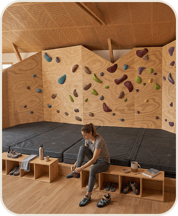 Group 658 Femme laçant ses chaussures d'escalade dans une salle d'escalade de bloc avec des murs en bois et des prises colorées.