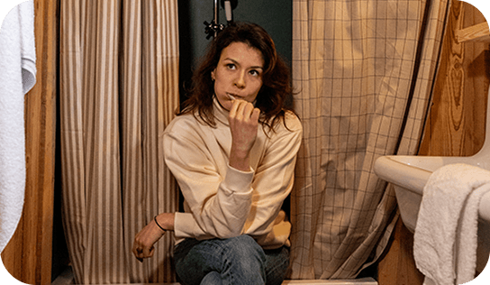 IMAGE-FAMILLE-3 Femme assise dans la salle de bain, l'air pensif. Couleurs neutres, rideau de douche rayé.