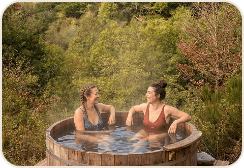 Rectangle 107 Deux femmes se détendent dans un bain à remous en bois à l'extérieur du « Camp de base », entourées d'une végétation luxuriante.